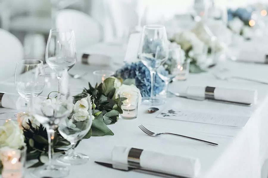 Special-Event-Insurance-Wedding-Table-Decoration-with-Floral-Garland-and-Blue-Flowers-Between-Glasses-on-a-White-Tablecloth